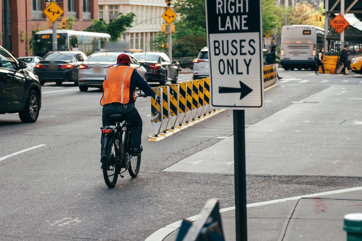 A cyclist in an orange vest riding along a city street next to a Buses Only sign and lane barriers