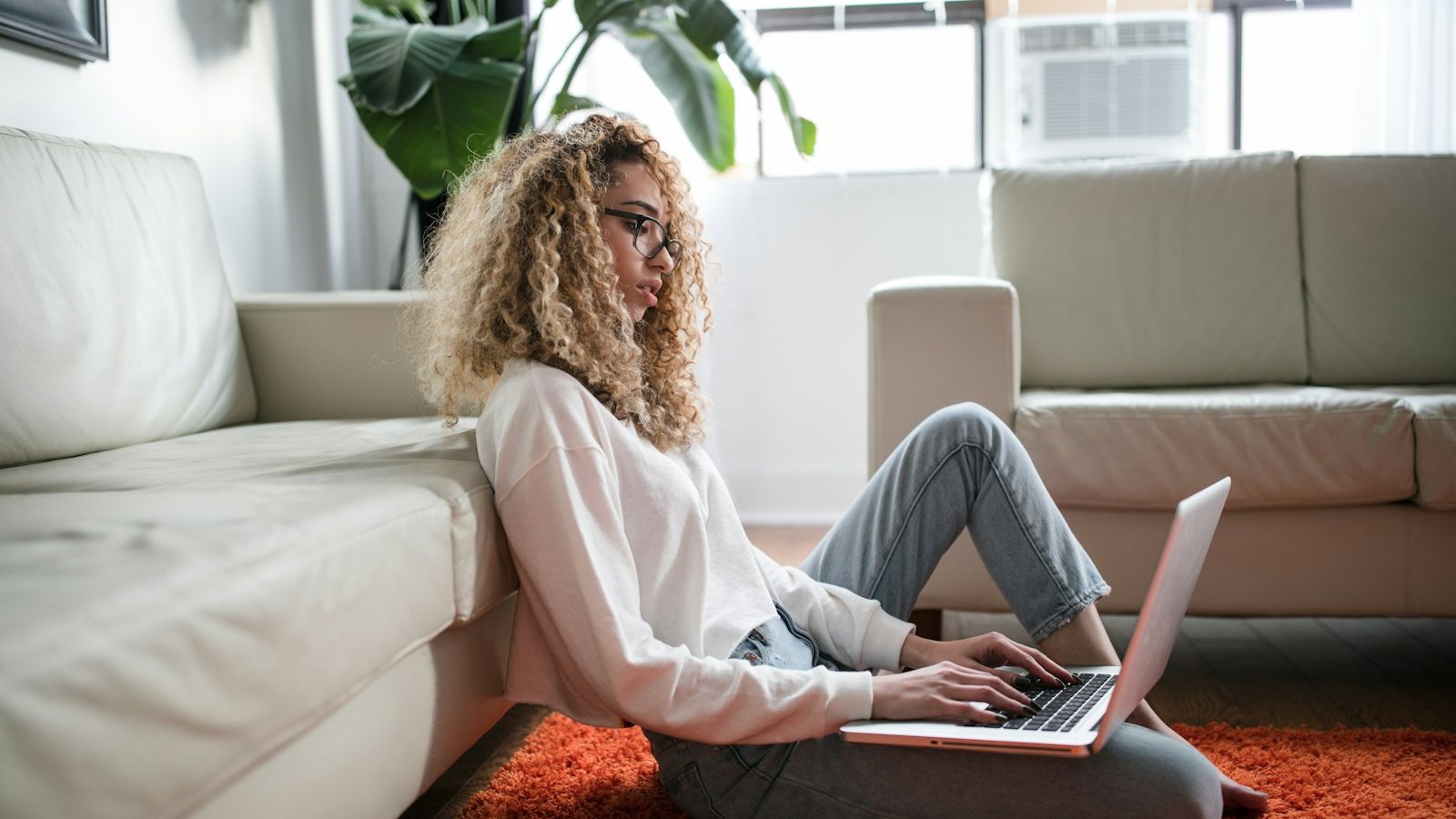 A woman with curly hair using a laptop while sitting on a rug by her sofa